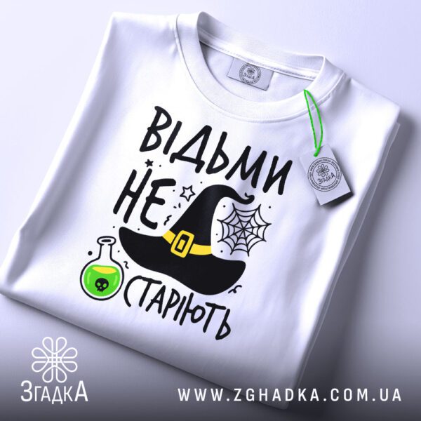 Складена біла футболка з принтом "відьми не старіють" та етикеткою "Згадка".
