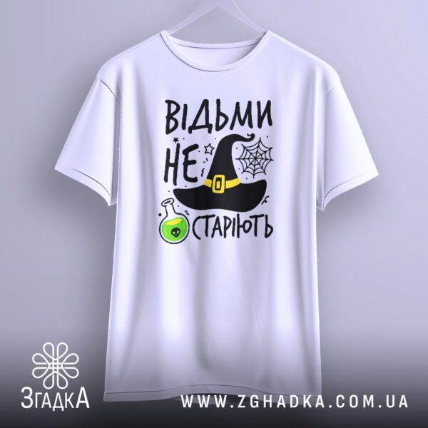 Біла футболка з принтом "відьми не старіють", що висить на білих плічках, демонструючи дизайн.