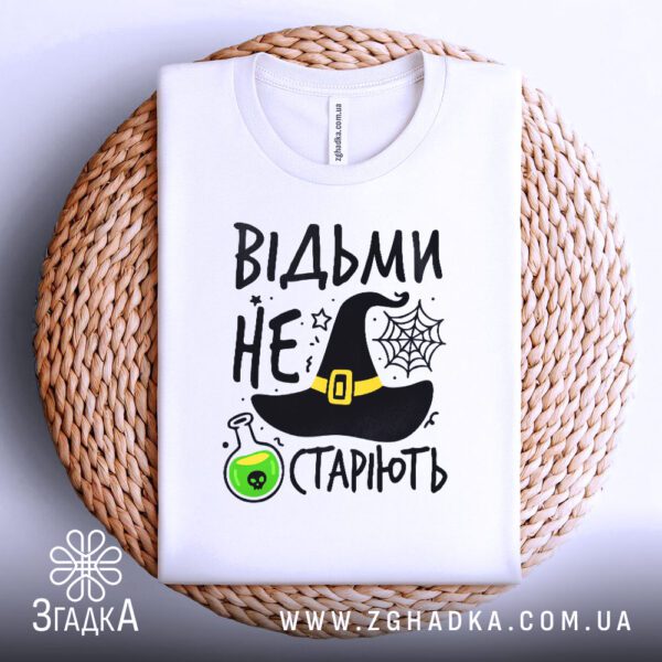 Біла футболка з принтом "відьми не старіють", акуратно складена на круглій дерев'яній поверхні.