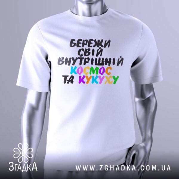 Біла футболка з яскравим, багатобарвним графічним принтом у центрі. Принт виконаний у стилі мультяшного або коміксного зображення, що нагадує галактику або космос з зірками та планетами. У центрі композиції великими літерами написано фразу: "БЕРЕЖИ СВІЙ ВНУТРІШНІЙ КОСМОС ТА КУКУХУ". Футболка на сірому манекені.