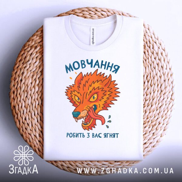Біла футболка з принтом "мовчання робить з вас ягнят", акуратно складена на круглому плетеному підносі.
