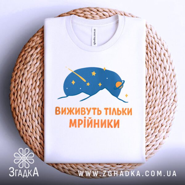 Біла футболка з принтом "виживуть тільки мрійники", акуратно складена на круглій дерев'яній поверхні.