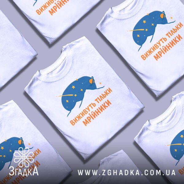 Кілька білих футболок з принтом "виживуть тільки мрійники", складені стопкою або розкладені поруч на світлому фоні.