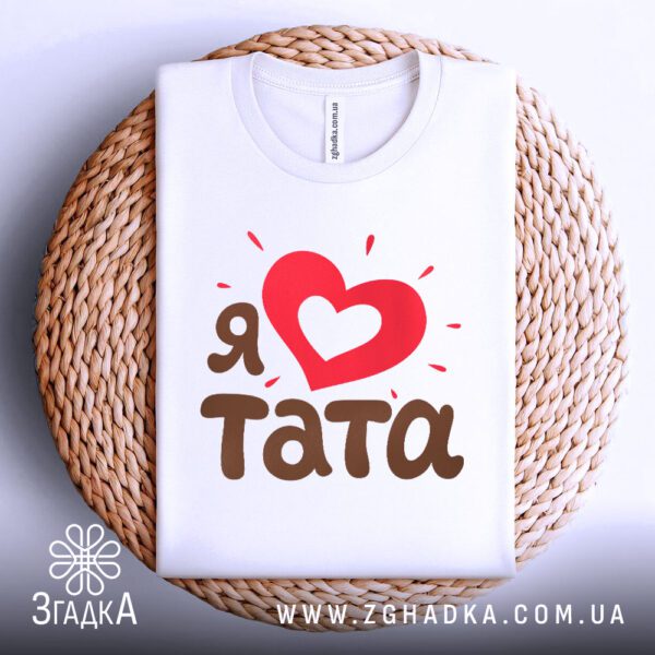 Біла футболка з принтом "Я ♥ тата", акуратно складена на круглій дерев'яній поверхні.