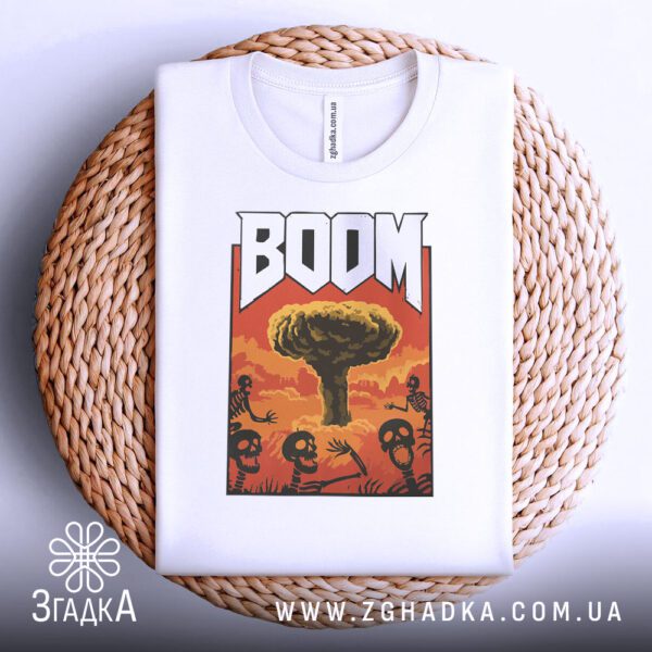 Біла футболка з принтом "ядерний вибух BOOM", акуратно складена на круглій дерев'яній поверхні.