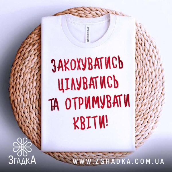 Біла футболка з принтом "любовний посил", акуратно складена на круглій дерев'яній поверхні.