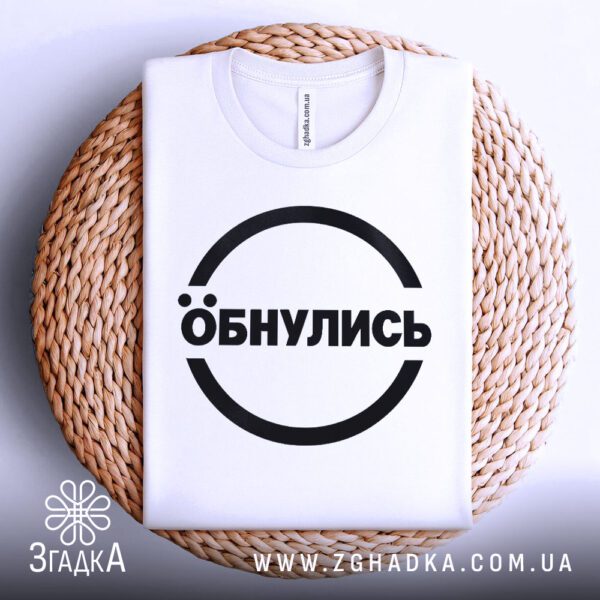 Біла футболка з принтом "Обнулись", акуратно складена на круглій дерев'яній поверхні.