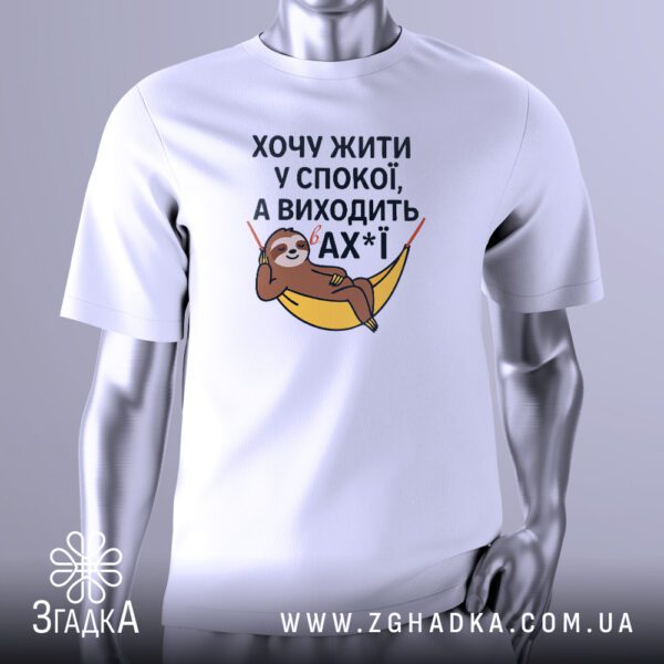 Біла футболка з графічним принтом у центрі. На принті зображений стилізований лінивець, що лежить у гамаку. Над ним і під ним розташований жартівливий текст українською мовою: "Хочу жити у спокої, а виходить в ах*ї". Футболка на сірому манекені.