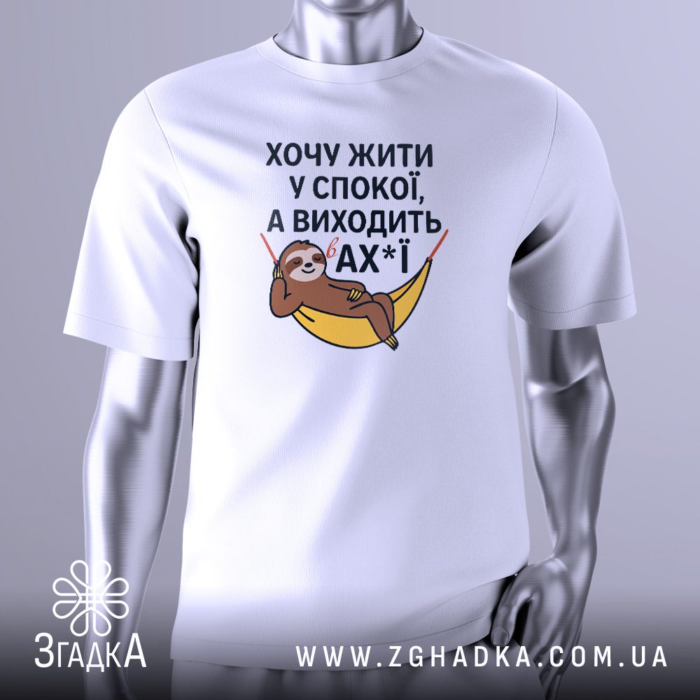 Біла футболка з графічним принтом у центрі. На принті зображений стилізований лінивець, що лежить у гамаку. Над ним і під ним розташований жартівливий текст українською мовою: "Хочу жити у спокої, а виходить в ах*ї". Футболка на сірому манекені.
