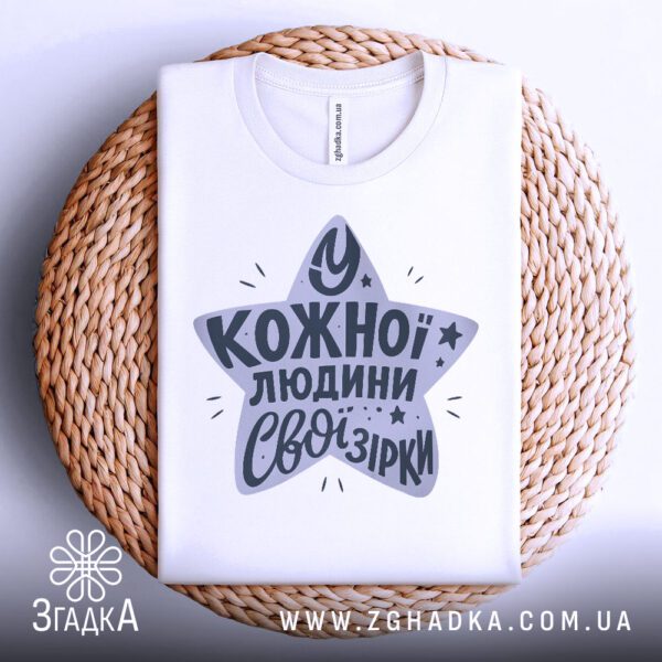 Біла футболка з принтом "зірки всередині нас", акуратно складена на круглій дерев'яній поверхні.