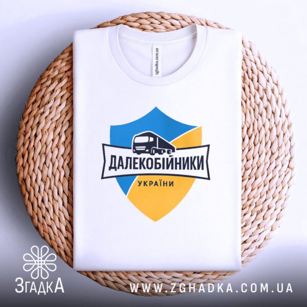 Біла футболка з принтом "далекобійники України патріотична", акуратно складена на круглій дерев'яній поверхні.