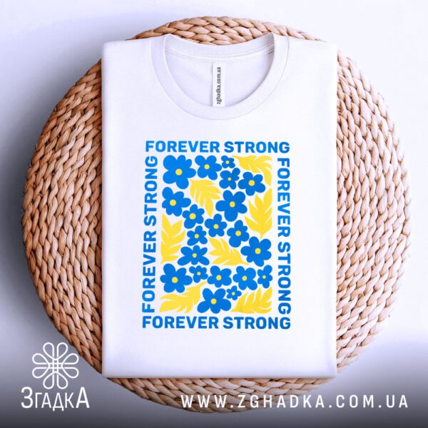 Біла футболка з принтом "forever strong Україна", акуратно складена на круглій дерев'яній поверхні.