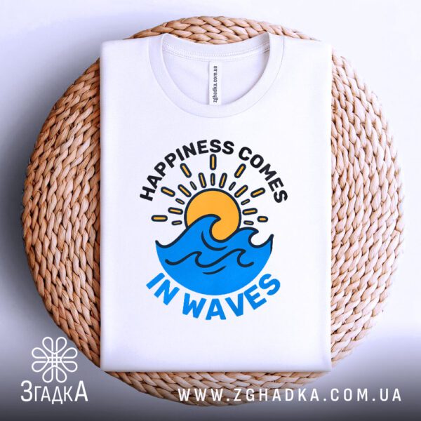 Біла футболка з принтом "happiness comes waves", акуратно складена на круглій дерев'яній поверхні.