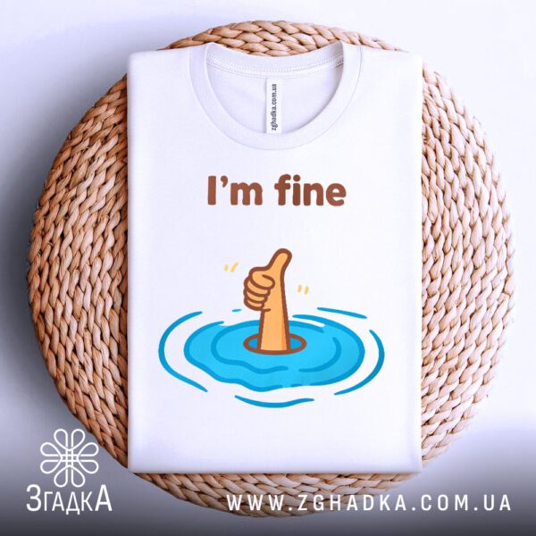 Біла футболка з принтом "I'm fine гумор", акуратно складена на круглій дерев'яній поверхні.