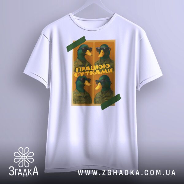Біла футболка з принтом "качки працюю сутками", що висить на вішаку, демонструючи дизайн.