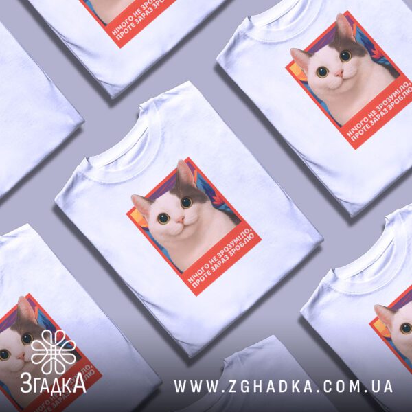 Біла футболка з принтом «котик з підписом», складена на поверхні.