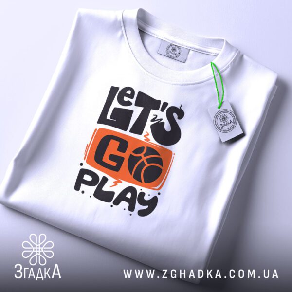 Складена біла футболка з принтом "let’s go play" та етикеткою "Згадка".