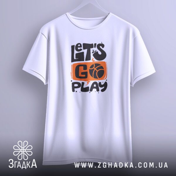 Біла футболка з принтом "let’s go play", що висить на білих плічках, демонструючи дизайн.