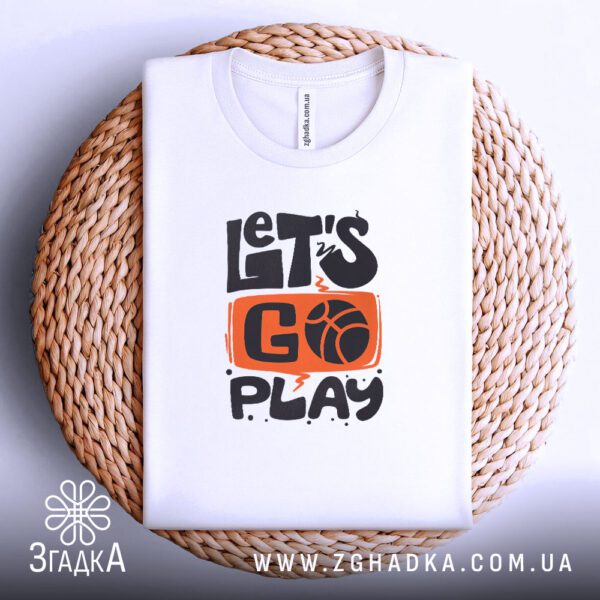 Біла футболка з принтом "let’s go play", акуратно складена на круглій дерев'яній поверхні.