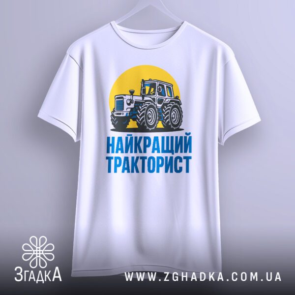 Біла футболка з принтом "найкращий тракторист подарунок", що висить на вішаку, демонструючи дизайн.