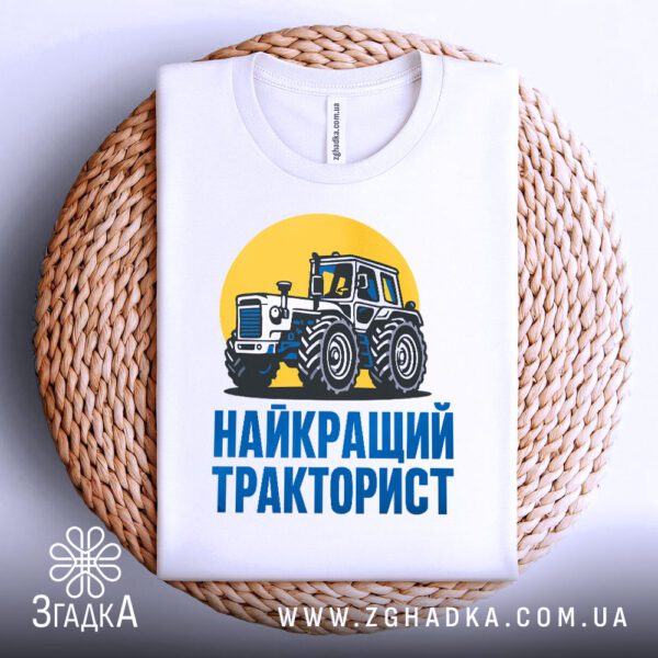Біла футболка з принтом "найкращий тракторист подарунок", складена на світлій поверхні.