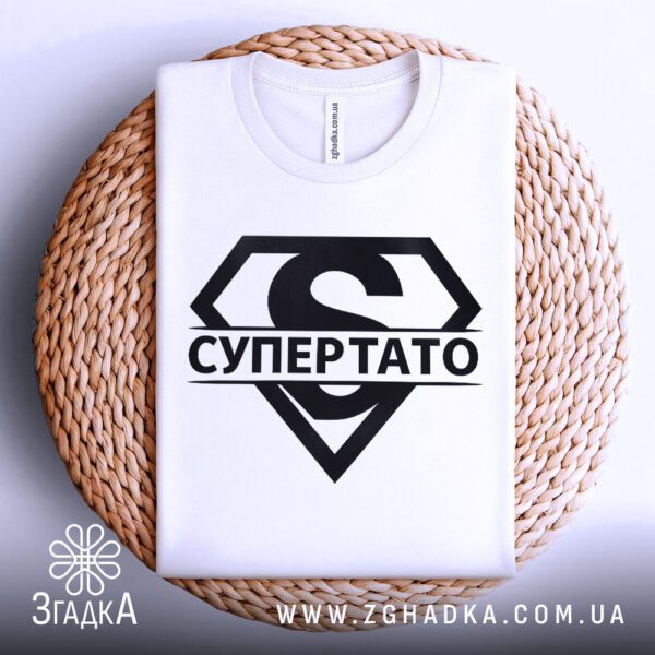 Біла футболка з принтом «супергерой тато», що висить на вішаку.