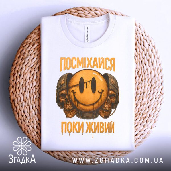 Біла футболка з принтом «посміхайся поки живий», що висить на вішаку.