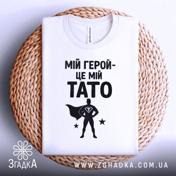 Біла футболка з принтом «тато завжди герой», що висить на вішаку.