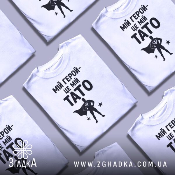 Біла футболка з принтом «тато завжди герой», складена на поверхні.