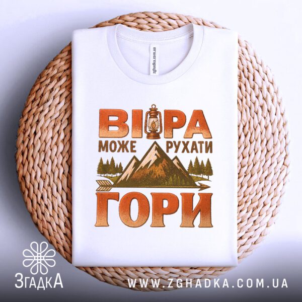 Біла футболка з принтом «віра рухає гори», що висить на вішаку.
