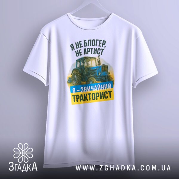 Біла футболка з принтом "я звичайний тракторист", що висить на вішаку, демонструючи дизайн.