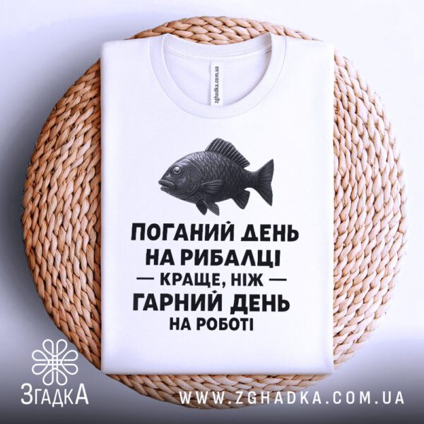 Біла футболка з написом "з написом рибалка", складена на світлій поверхні.