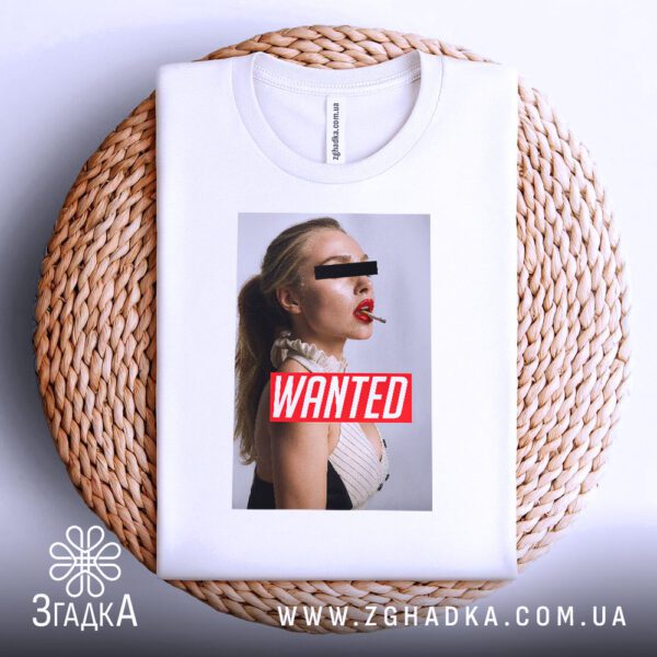 Біла футболка з принтом "з написом Wanted", акуратно складена на круглій дерев'яній поверхні.