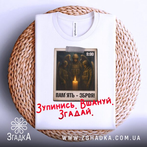 Біла футболка з принтом «Зупинись Вшануй Згадай», що висить на вішаку.