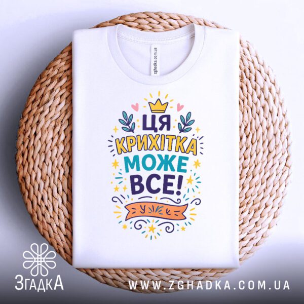Біла футболка з написом "Ця крихітка може все!", складена.