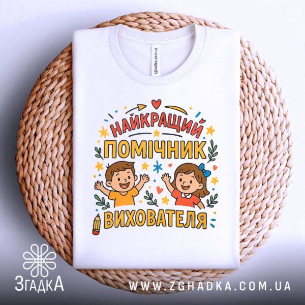 Футболка найкращий помічник вихователя на білому фоні, рівно складена.