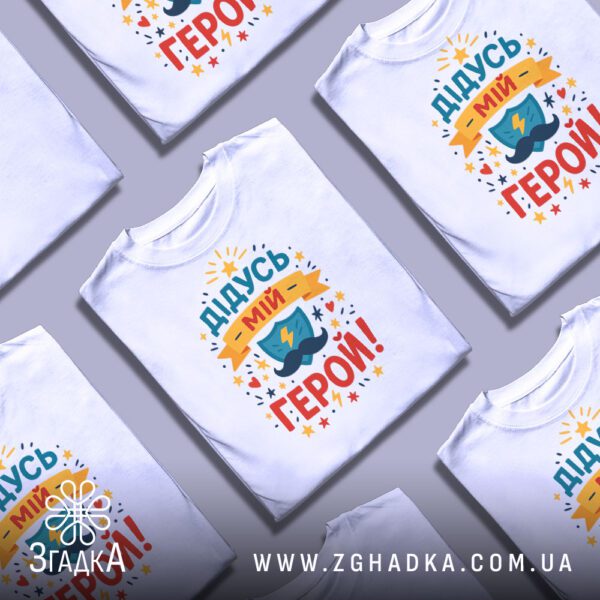 Біла футболка з принтом "Дідусь мій герой!" на вішалці.
