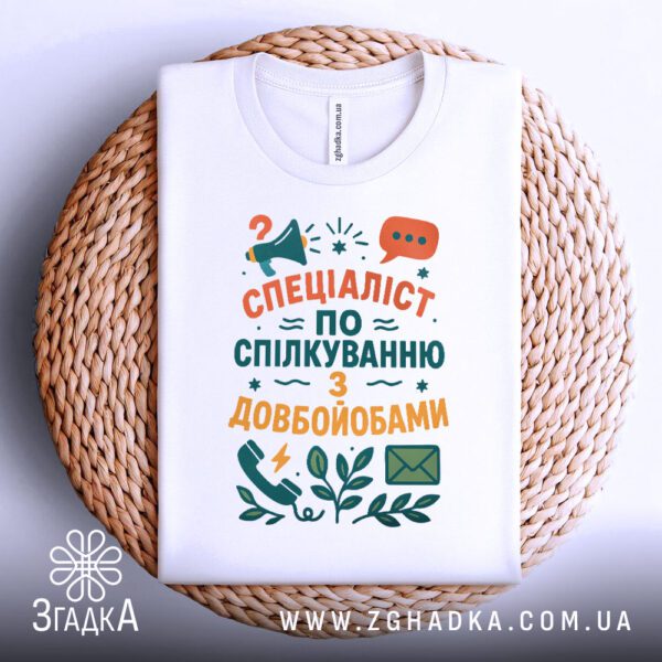 Біла футболка з принтом "Спеціаліст по спілкуванню з довбойобами", складена.