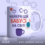 Чашка бабусі день народження. Чашка білого кольору з принтом, що містить напис 'НАЙКРАЩИЙ БАБУСІ НА СВІТІ' у червоному та фіолетовому кольорах, а також ілюстрації у вигляді сердець, окулярів та клубка пряжі. Ручка чашки з класичним дизайном, край рівний, поверхня глянцева. Фон світло-фіолетовий з малюнками окулярів.