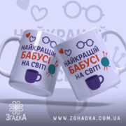 Чашка бабусі день народження. Дві білосніжні чашки з принтом, що містить напис 'НАЙКРАЩИЙ БАБУСІ НА СВІТІ', розташовані під кутом один до одного. Принт містить червоні серця, фіолетові окуляри та клубок пряжі. Ручки чашок класичні, краї рівні, поверхня глянцева. Фон світло-фіолетовий з малюнками окулярів.