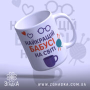 Чашка бабусі день народження. Чашка білого кольору з принтом, що містить напис 'НАЙКРАЩИЙ БАБУСІ НА СВІТІ' у червоному та фіолетовому кольорах, а також ілюстрації у вигляді сердець, окулярів та клубка пряжі. Ручка чашки з класичним дизайном, край рівний, поверхня глянцева. Фон світло-фіолетовий з малюнками окулярів.