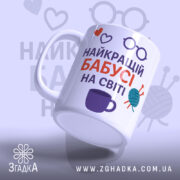 Чашка бабусі день народження. Чашка білого кольору з принтом, що містить напис 'НАЙКРАЩИЙ БАБУСІ НА СВІТІ' у червоному та фіолетовому кольорах, а також ілюстрації у вигляді сердець, окулярів та клубка пряжі. Ручка чашки з класичним дизайном, край рівний, поверхня глянцева. Фон світло-фіолетовий з малюнками окулярів.