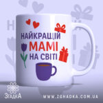 Чашка мамі день народження, біла з кольоровим принтом, зображення з фронтального ракурсу, фон світло-фіолетовий, на чашці напис "НАЙКРАЩИЙ МАМІ НА СВІТІ" червоним і фіолетовим кольором, з малюнками сердець, подарунків і тюльпана, ручка класична, край гладкий, глянцеве покриття.