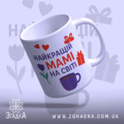 Чашка мамі день народження, біла з кольоровим принтом, зображення з верхнього ракурсу, фон світло-фіолетовий, на чашці напис "НАЙКРАЩИЙ МАМІ НА СВІТІ" червоним і фіолетовим кольором, з малюнками сердець, подарунків і тюльпана, ручка класична, край гладкий, глянцеве покриття.