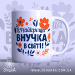 Чашка найкраща внучка подарунок. Чашка білого кольору з яскравим принтом, на якому зображені червоні та помаранчеві квіти, а також текст синього кольору. Ручка круглої форми, край рівний, поверхня глянцева. Фон ніжно-фіолетовий з абстрактними квітковими елементами.
