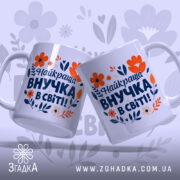 Чашка найкраща внучка подарунок. Дві чашки білого кольору, повернуті одна до одної, з яскравим принтом, на якому зображені червоні та помаранчеві квіти, а також текст синього кольору. Ручки круглої форми, краї рівні, поверхня глянцева. Фон ніжно-фіолетовий з абстрактними квітковими елементами.
