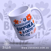 Чашка найкраща внучка подарунок. Чашка білого кольору з яскравим принтом, на якому зображені червоні та помаранчеві квіти, а також текст синього кольору. Ручка круглої форми, край рівний, поверхня глянцева. Чашка нахилена, фон ніжно-фіолетовий з абстрактними квітковими елементами.