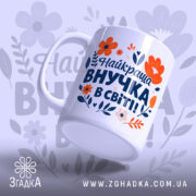 Чашка найкраща внучка подарунок. Чашка білого кольору з яскравим принтом, на якому зображені червоні та помаранчеві квіти, а також текст синього кольору. Ручка круглої форми, край рівний, поверхня глянцева. Чашка нахилена, фон ніжно-фіолетовий з абстрактними квітковими елементами.