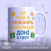 Чашка найкращій доні подарунокʼ Біла чашка з кольоровим принтом, на якому написано 'Ця чашка належить найкращій доні в світі'. Ручка чашки гладка, край рівний, фон ніжно-фіолетовий, з елементами жовтих зірочок, квітів та подарунків. Чашка має глянцеву поверхню.