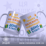Чашка найкращій доні подарунокʼ Дві білі чашки, розташовані під кутом, з кольоровим принтом, на якому написано 'Ця чашка належить найкращій доні в світі'. Ручки чашок гладкі, краї рівні, фон ніжно-фіолетовий з елементами жовтих зірочок, квітів та подарунків. Чашки мають глянцеву поверхню.